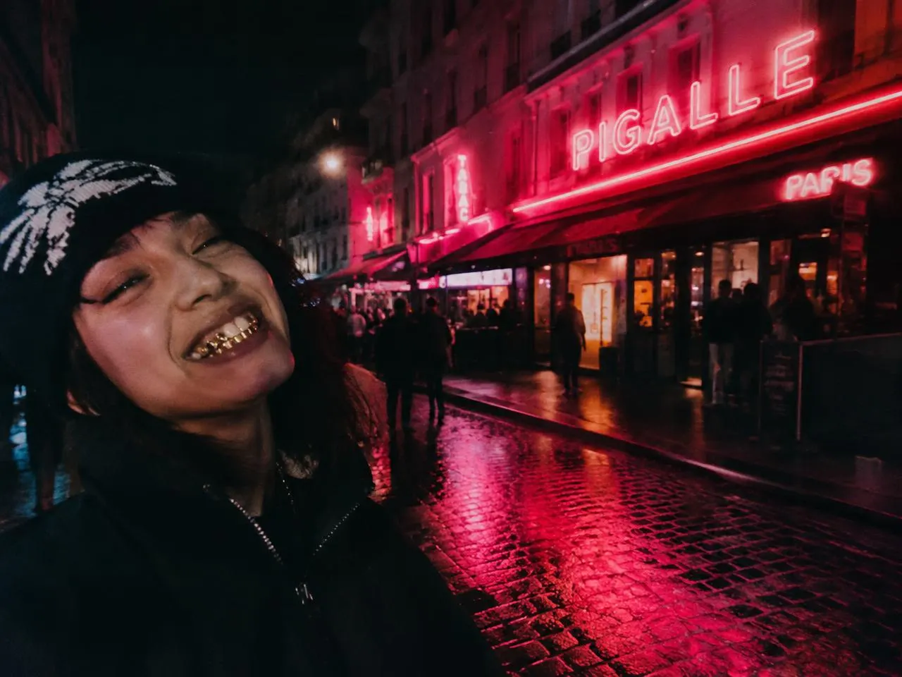 Woman laughing on a rainy Pigalle street at night showing gold grillz under red neon signs